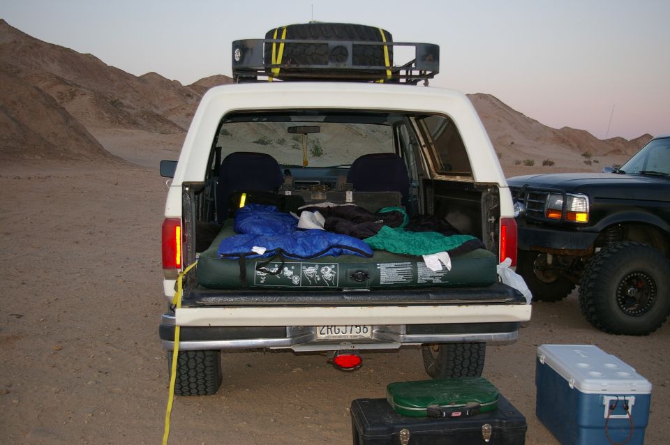 White Ford Bronco rear view with camping gear in the bed and a black Ford truck beside it