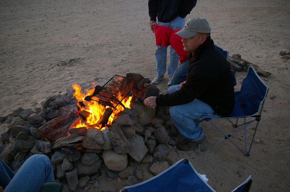 People sitting around a campfire on a sandy area, cooking food on a grill over the flames