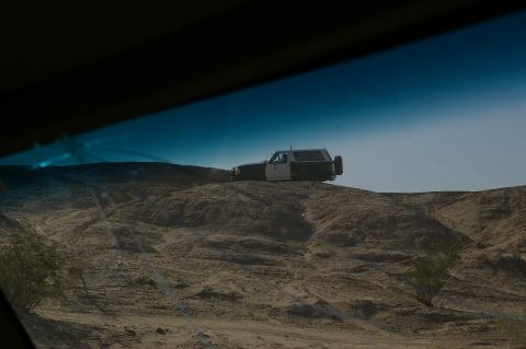 1986 Ford Bronco - A two-tone SUV is seen from inside a vehicle, parked on a barren, dusty landscape under a clear blue sky.