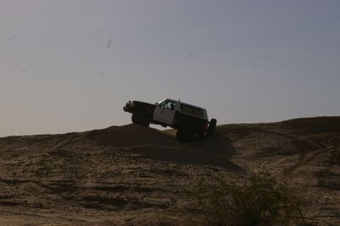 1986 Ford Bronco - A two-tone SUV navigates a sandy incline under a clear sky.