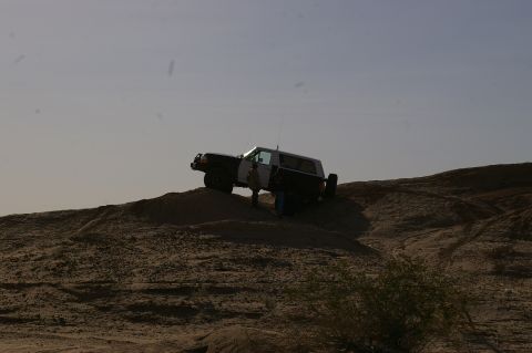 1986 Ford Bronco - A two-tone 1986 Ford Bronco with a camper shell navigates a sandy incline under a hazy sky.