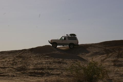 1986 Ford Bronco - White 1986 Ford Bronco navigates a sandy incline under a clear sky.