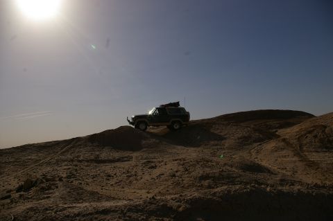 1986 Ford Bronco - A dark green Ford Bronco navigates a sandy terrain under a bright, hazy sky.