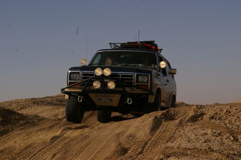 1986 Ford Bronco - 1986 Ford Bronco navigates sandy terrain under a clear sky, equipped with auxiliary lighting and a roof rack.