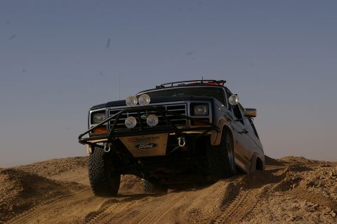 1986 Ford Bronco - This modified Ford Bronco navigates a sandy terrain under a clear blue sky.