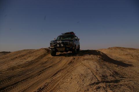 1986 Ford Bronco - A dark SUV navigates sandy terrain under a clear blue sky.