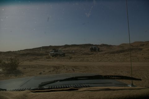 1986 Ford Bronco - Two SUVs traverse a sandy terrain under a vast blue sky, suggesting an off-road adventure.