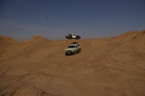 1986 Ford Bronco - Two SUVs navigate sandy terrain, one following the other up a dune.