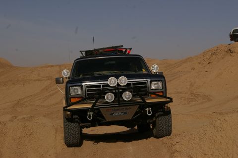 1986 Ford Bronco - A dark blue Ford Bronco navigates sandy terrain, equipped with auxiliary lights and a roof rack.