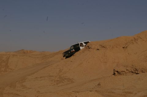 1986 Ford Bronco - A white and black SUV navigates a sandy terrain, climbing a large dune.