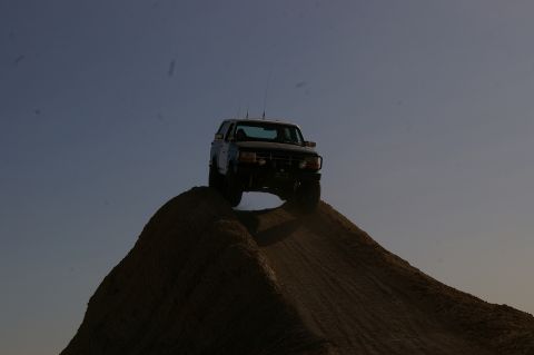 1986 Ford Bronco - A Ford Bronco is captured mid-climb on a steep dirt mound under a clear sky.