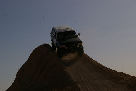 1986 Ford Bronco - SUV with a white camper shell navigates a steep dirt incline under a clear sky.