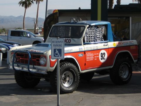 1996 Ford Bronco - This modified Ford Bronco, likely from the 1970s, is parked in a lot with other vehicles and palm trees.