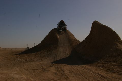 1986 Ford Bronco - A white SUV navigates a steep dirt incline, its roof loaded with gear, under a clear blue sky.