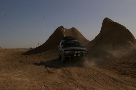 1986 Ford Bronco - A Ford Bronco kicks up dust as it navigates a desert trail near unusual geological formations.