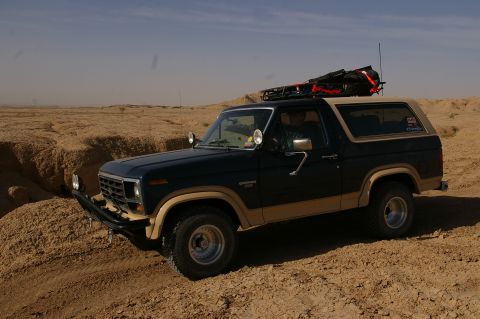 1986 Ford Bronco - A Ford Bronco navigates a sandy terrain under a clear sky, its roof rack packed for an adventure.