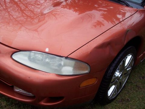 1995 Mitsubishi Eclipse - The front corner of an orange car shows a dent on the fender and a clear headlight.