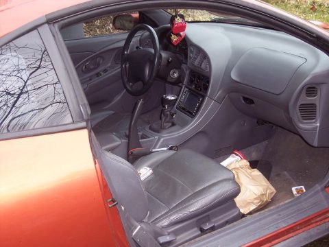 1995 Mitsubishi Eclipse - The interior of a 1995 Mitsubishi Eclipse is visible, including the dashboard, seats, and a paper bag on the floor.