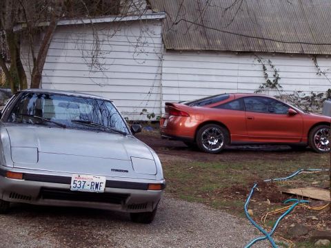 1995 Mitsubishi Eclipse - Two sports cars, a silver Mazda RX-7 and an orange Mitsubishi Eclipse, are parked outside a building.