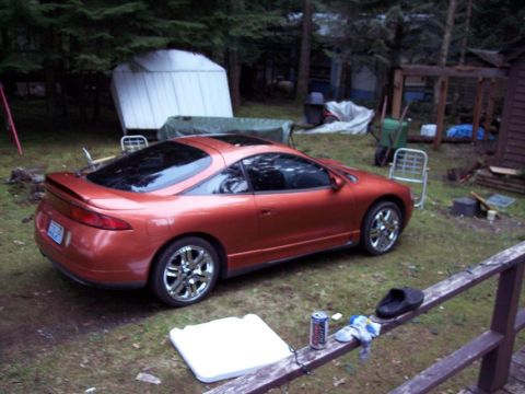 1995 Mitsubishi Eclipse - This orange sports coupe is parked in a wooded area next to a shed and some lawn equipment.