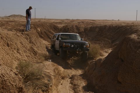 1986 Ford Bronco - A two-tone SUV navigates a challenging off-road path in a desert landscape.