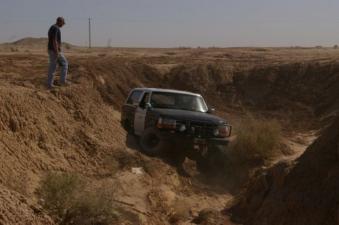 1986 Ford Bronco - A man observes a Ford Bronco navigating a steep, dry, dirt gully in a desert landscape.