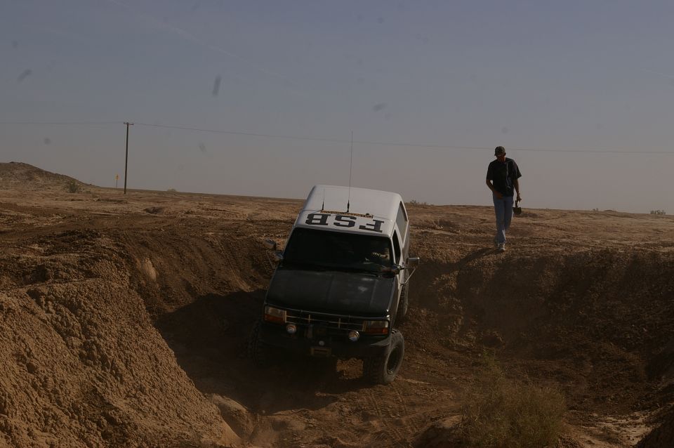 1986 Ford Bronco driving down a dirt embankment with a person walking on the ridge above
