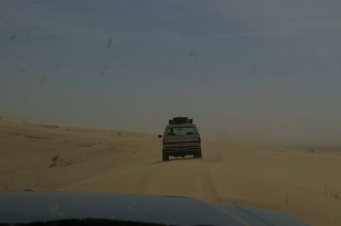 1986 Ford Bronco - A white SUV navigates a dusty desert track, kicking up sand as it drives away.