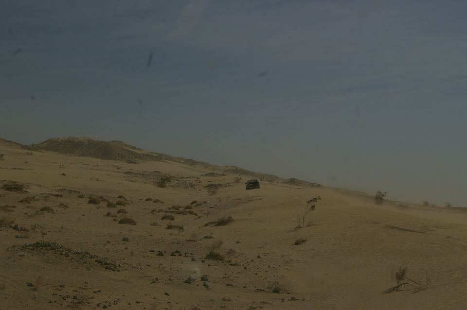 Desert terrain with two dark-colored vehicles driving on a sandy ridge under a hazy blue sky