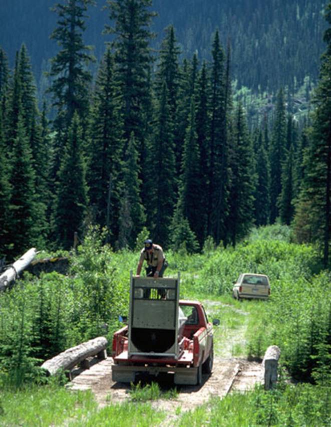 red pickup truck with a man standing on a metal frame in the bed, another vehicle in background on dirt road