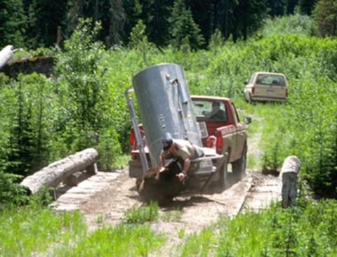 1974 Ford Ranchero - A red pickup truck navigates a rough terrain, carrying a large cylindrical tank, with another vehicle visible in the distance.