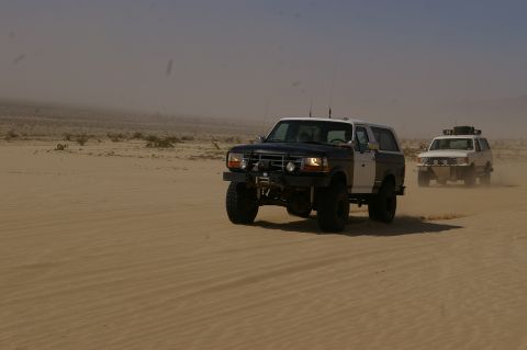 1986 Ford Bronco - Two lifted Ford Broncos kick up dust while driving through a desert environment.
