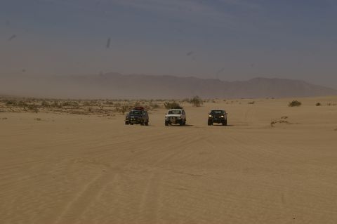 1986 Ford Bronco - Three SUVs traverse a desert landscape, kicking up dust.