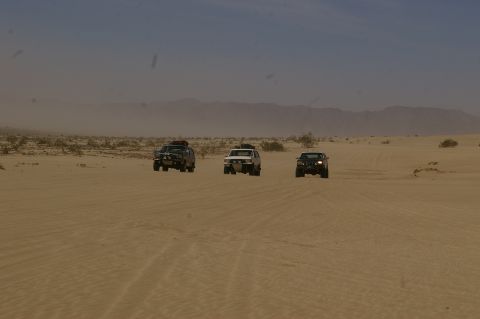 1986 Ford Bronco - A convoy of three SUVs traverses a sandy desert terrain under a hazy sky.