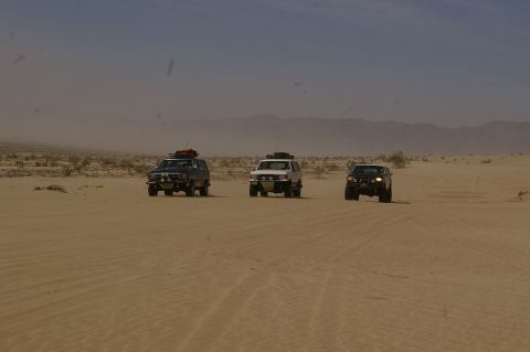 1986 Ford Bronco - Three SUVs traverse a dusty desert terrain, kicking up sand as they drive towards the horizon.