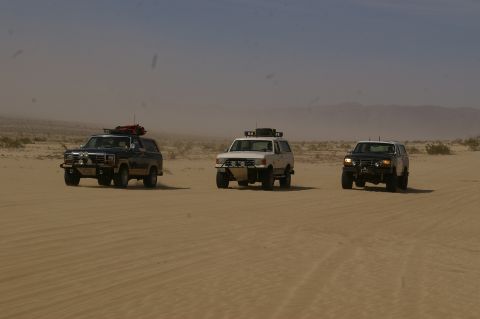 1986 Ford Bronco - Three SUVs traverse a sandy desert terrain under a hazy sky. The lead dark blue SUV resembles an early 1980s model.