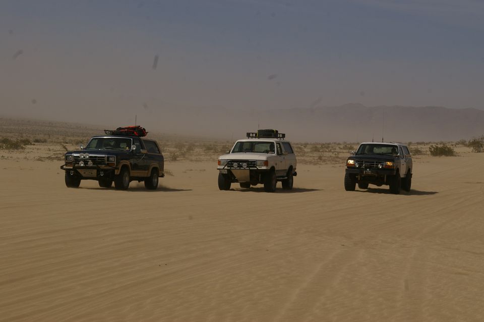 Three SUVs driving in a desert, one is dark blue, one is white, and one is black. The dark blue SUV appears to be from the early 1980s