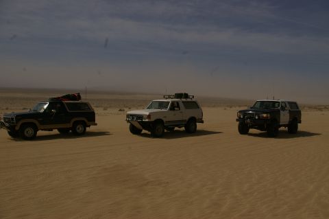 1986 Ford Bronco - Three SUVs traverse a sandy desert terrain, kicking up dust under a bright sky.