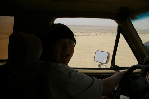 1986 Ford Bronco - A person drives a vehicle through a dusty desert environment, looking back at the camera.
