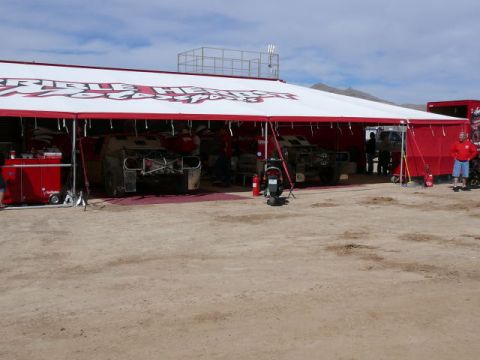 1988 Ford Bronco - A large red-and-white race team awning pitched on desert dirt shelters two off-road trucks up on stands with wheels removed and tube chassi…