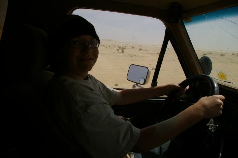 1986 Ford Bronco - A young person drives through a dusty, arid terrain under a hazy sky.
