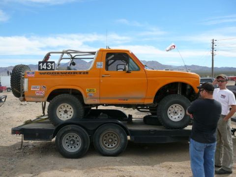 1988 Ford Bronco - Passenger-side profile of an orange full-size Ford Bronco desert race truck strapped to a flatbed tandem-axle trailer in the desert, with r…