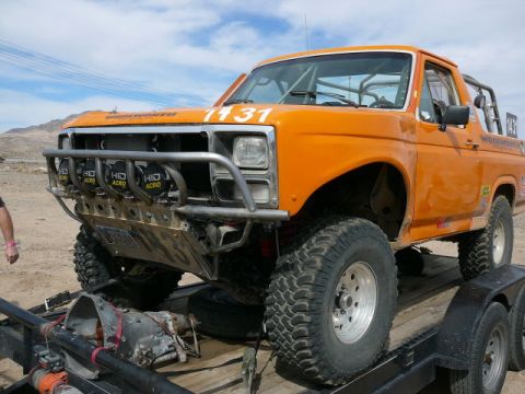 1988 Ford Bronco - Orange full-size Ford Bronco desert race truck with race number 1131 on the windshield, strapped to a wood-deck trailer in the desert. It w…