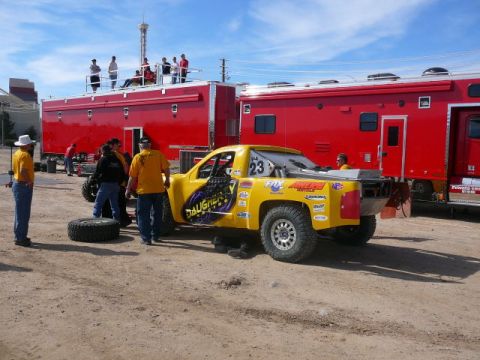 1988 Ford Bronco - Yellow and purple off-road race truck numbered 23 parked on dirt in a desert pit area, seen from the rear three-quarter with a flat aluminu…