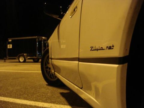 1991 Nissan 240SX - A white car is parked next to a black trailer at night. The car has a website sticker on its side.