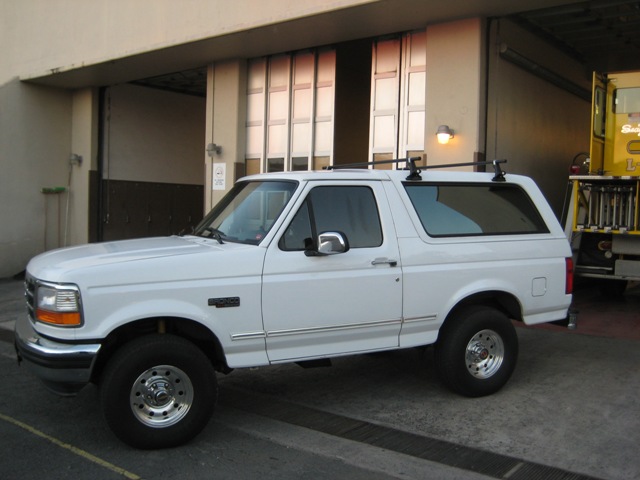 White Ford Bronco, 1992 model year, parked outside a building with a yellow truck in the background