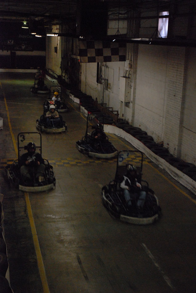 Go-karts racing indoors on a track with a checkered flag overhead. Some karts have numbers