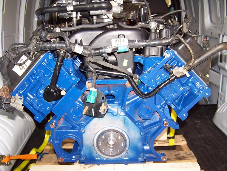 Blue painted V8 engine block with wiring harnesses and hoses attached, sitting on a wooden pallet inside a van