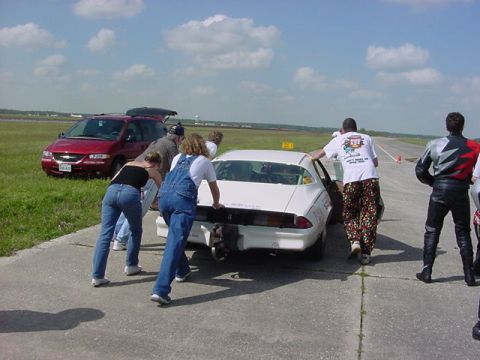 1979 Ford Bronco - People push a white coupe on a tarmac, with a red minivan parked nearby under a cloudy sky.