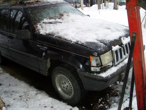 2000 Ford E350 - A black Jeep Grand Cherokee sits partially covered in snow, showing damage to its front end.